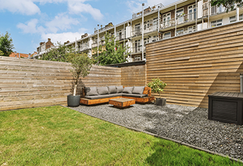 The image shows a beautifully remodeled backyard featuring a paved patio with a custom-built wooden seating area, a wooden coffee table, and a wooden fence. The space is enhanced with potted plants, creating a cozy and inviting outdoor living area.